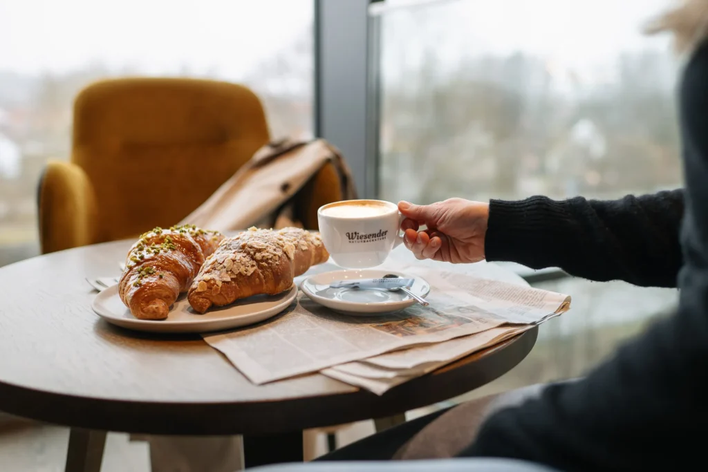 Eine Person sitzt an einem runden Tisch in Freising mit zwei Croissants aus der Naturbackstube Wiesender auf einem Teller, einer Tasse Kaffee und einer Zeitung.