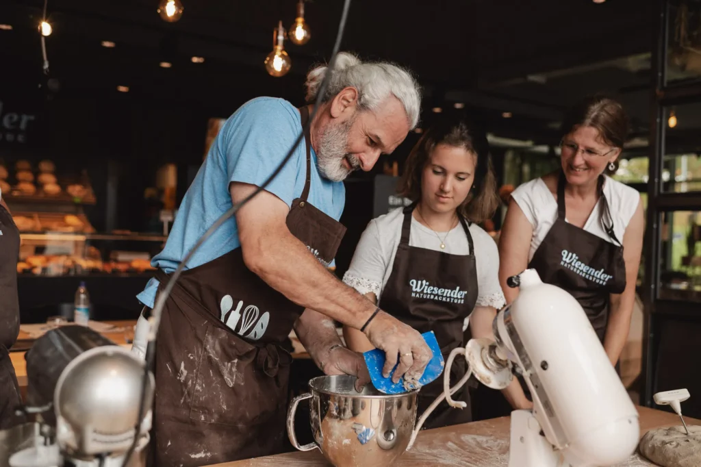 In der Bäckerei Wiesender in Pfaffenhofen arbeiten ein älterer Mann und zwei Frauen in braunen Schürzen gemeinsam in der Backstube. Der Mann, Karl Wiesender, kratzt Teig aus einer Rührschüssel, während die anderen lächelnd zusehen, umgeben von Backgeräten und frischen Zutaten.