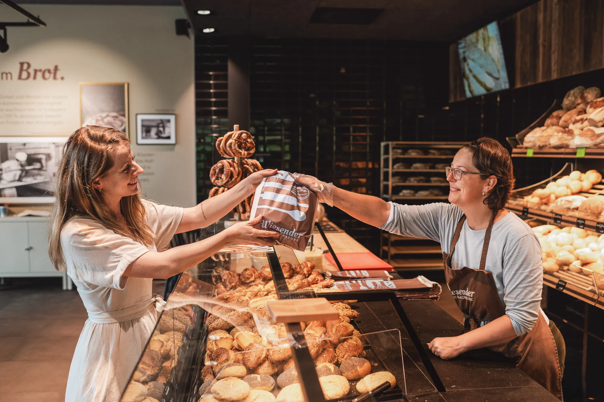 Eine lächelnde Frau hinter der Theke der Bäckerei Wiesender reicht einer Kundin eine Tüte mit Backwaren. Die Naturbackstube ist mit Brot und Gebäck gefüllt, im Hintergrund steht noch mehr frisches Brot in den Regalen.