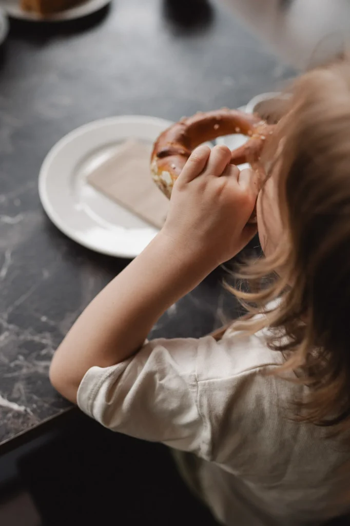 Ein kleines Kind mit langen Haaren sitzt an einem Tisch, hält eine Breze der Bäckerei Wiesender in der Hand und isst sie auf einem weißen Teller mit einer Serviette. Die Szene ist von oben und hinter dem Kind aufgenommen.