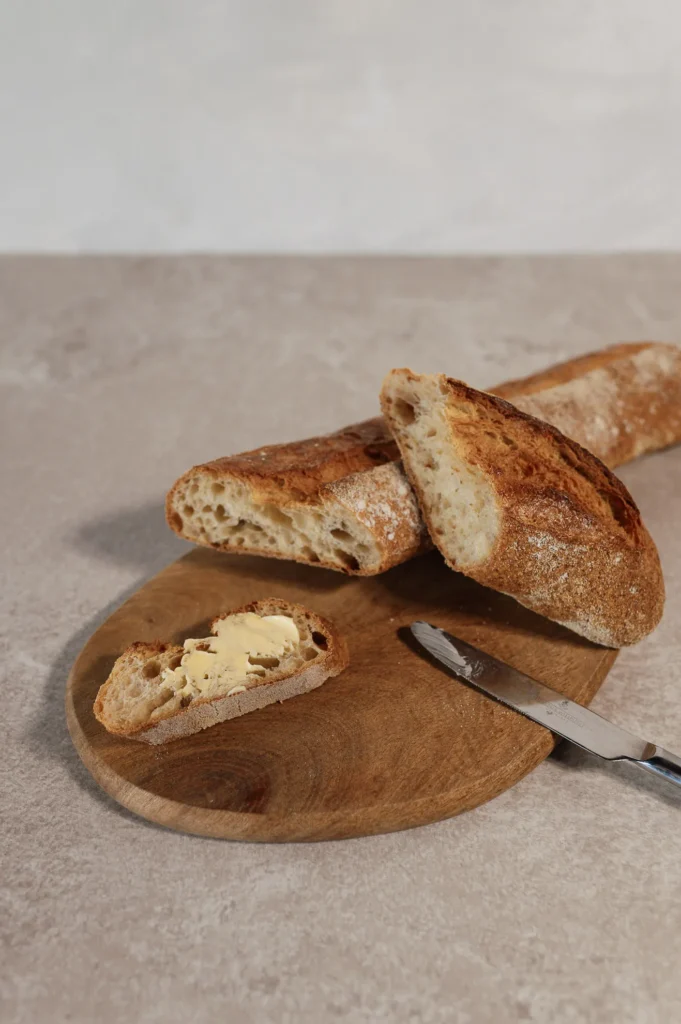 Ein aufgeschnittenes Baguette der Bäckerei Wiesender auf einem Holzbrett, ein Stück davon mit Butter bestrichen. Neben dem Brot liegt auf einer hellen Fläche ein Buttermesser.