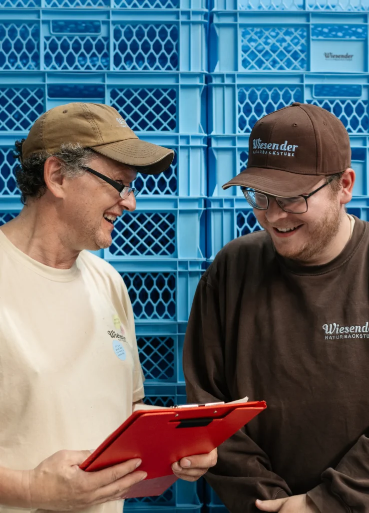 Zwei Männer mit Wiesender-Cpas und -Hemden lächeln und schauen auf ein rotes Klemmbrett. Sie stehen vor einem Stapel blauer Plastikkisten am Standort der Bäckerei Wiesender in Pfaffenhofen.