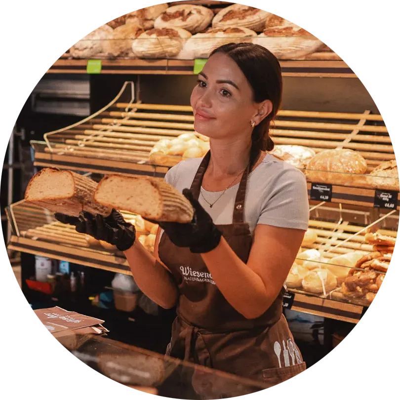Eine Frau mit Schürze und Handschuhen steht in der Bäckerei Wiesender und hält zwei Hälften eines rustikalen Brotlaibs in der Hand. Im Hintergrund füllen Regale mit verschiedenen Brotsorten die Bäckerei in Pfaffenhofen oder Freising.