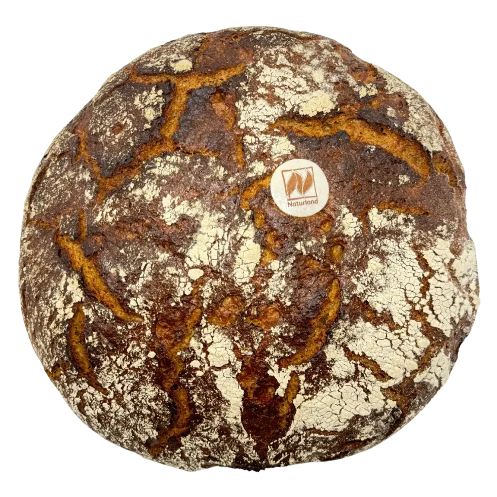 Rundes, rustikales, dunkelbraunes Brot mit rissiger, mehlbestäubter Kruste und einem kleinen, runden Naturland Etikett auf der Oberseite - mit Sorgfalt in der Naturbackstube in Freising hergestellt.