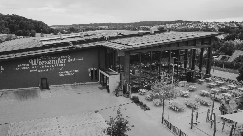 Ein großes, modernes Bäckereigebäude - die Bäckerei Wiesender - mit raumhohen Fenstern und Sitzplätzen im Freien, umgeben von Hügeln und Häusern. In Pfaffenhofen gelegen, zeigt das Äußere stolz seinen Namen.
