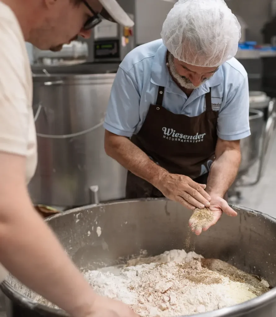In einer Naturbackstube in der Nähe von Pfaffenhofen bereiten zwei Männer Zutaten in der Bäckerei Wiesender vor. Der eine, der ein Haarnetz und eine Schürze trägt, streut Samen oder Körner aus seiner Hand in eine große Metallschüssel mit Mehl, während der andere genau beobachtet.