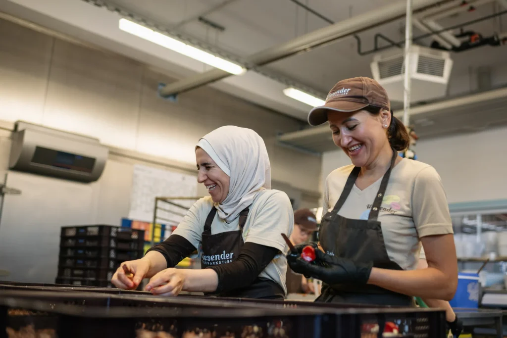 Zwei Frauen mit Schürzen und Mützen arbeiten zusammen und lächeln, während sie in der Naturbackstube der Bäckerei Wiesender in Freising einpacken. Eine Frau trägt einen weißen Hidschab, die andere eine braune Mütze und Handschuhe.