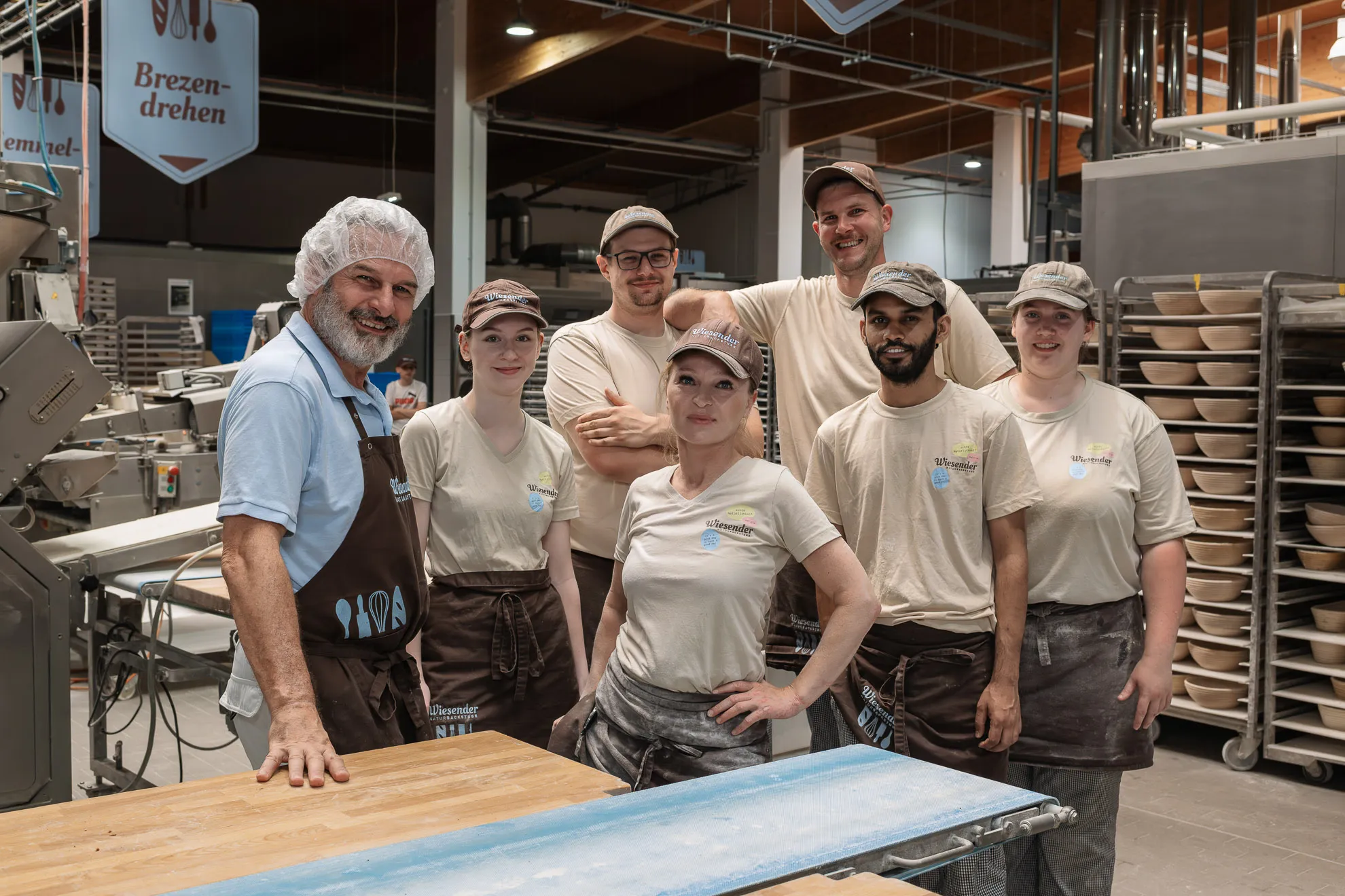 Sechs Bäckereiangestellte und der Chef Karl Wiesender in Arbeitskleidung und mit Schürzen stehen lächelnd in der Bäckerei Wiesender Naturbackstube in Pfaffenhofen, umgeben von Regalen mit Brot und Backgeräten.