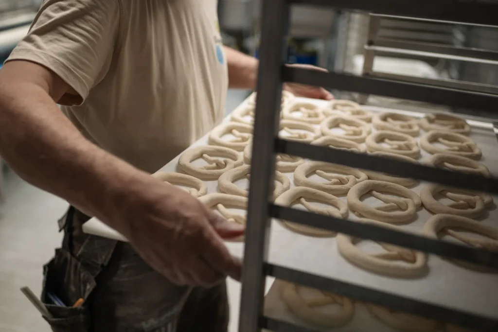In der Naturbackstube der Bäckerei Wiesender in Freising legt eine Person ein Tablett mit rohem, gedrehtem Brezelteig auf ein Metallgestell in der Backstube.
