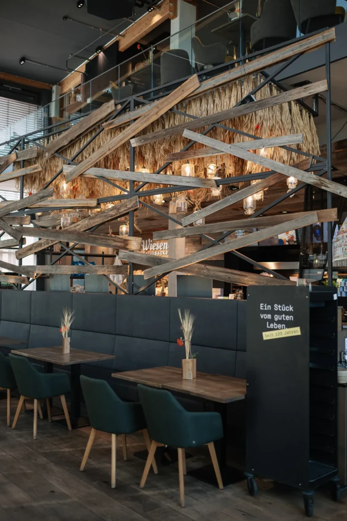 Ein modernes Café in Pfaffenhofen mit schwarzen Sitzpolstern, grünen Stühlen, Holztischen und rustikalen Balken, die mit hängenden Gläsern dekoriert sind.