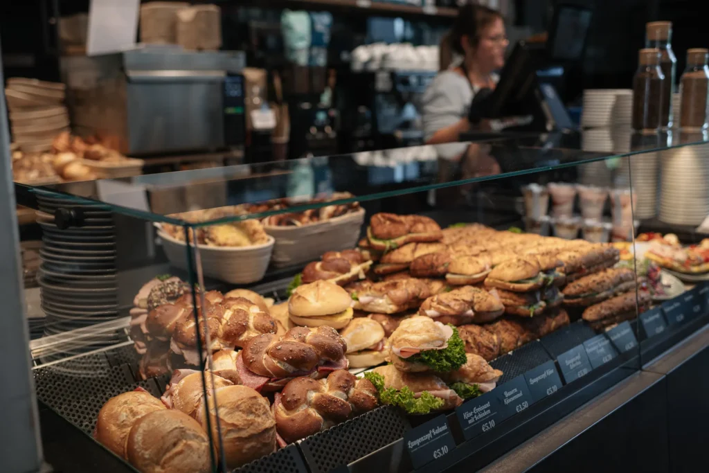 In der Naturbackstube in Freising ist eine Bäckereivitrine mit verschiedenen belegten Brötchen und Gebäck gefüllt, hinter dem Tresen steht eine Mitarbeiterin, im Hintergrund die Kasse.