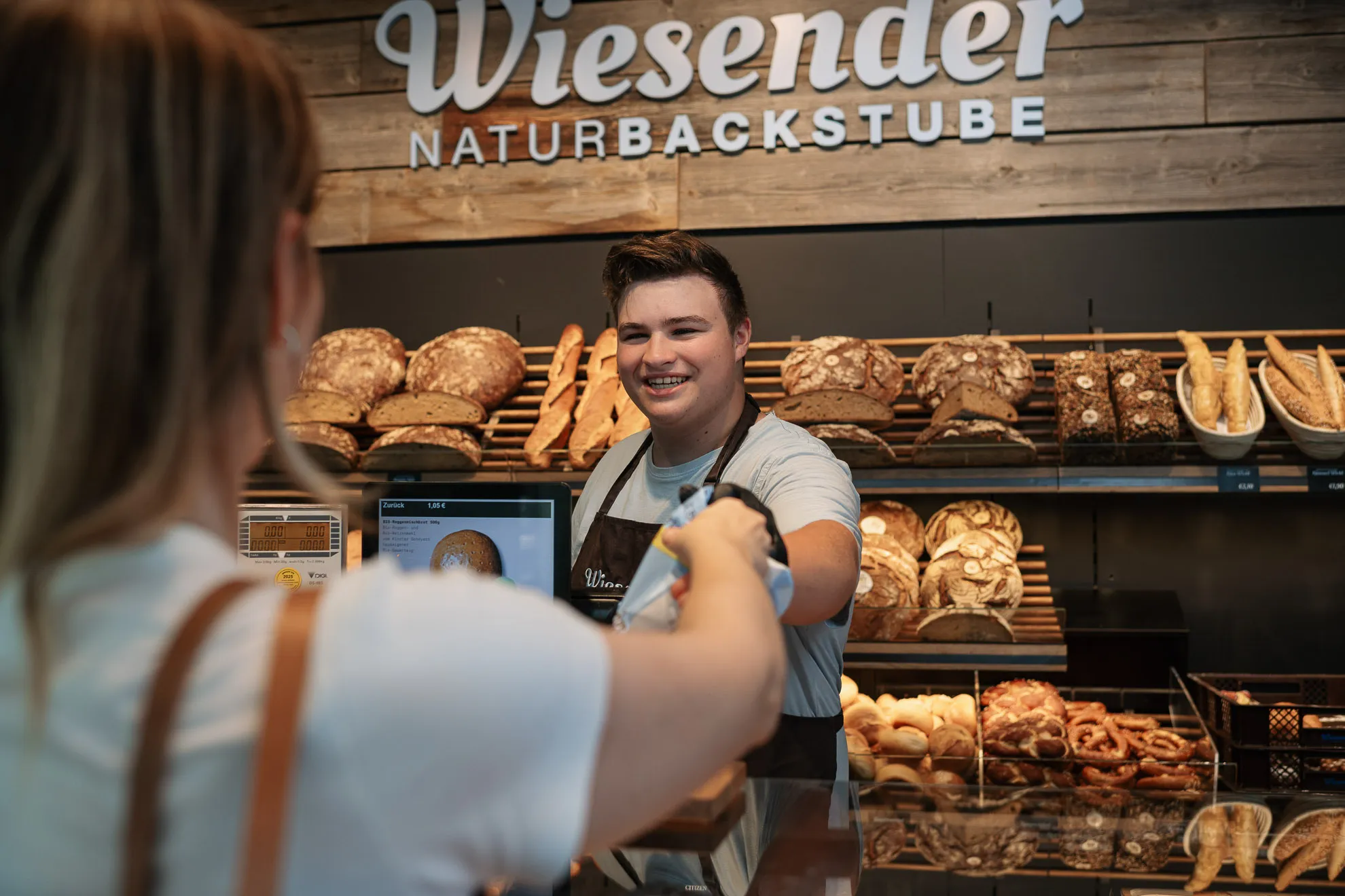 Ein lächelnder Bäckereimitarbeiter der Bäckerei Wiesender überreicht einem Kunden an der Theke einen Einkauf. Dahinter stehen Regale mit verschiedenen Broten und Brezen.
