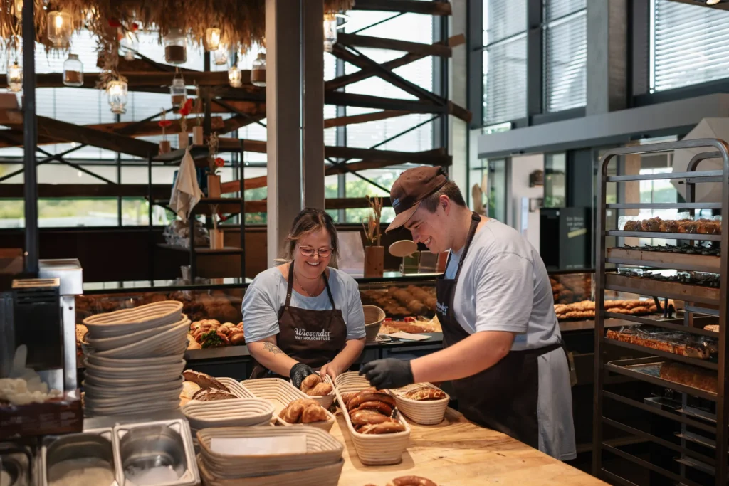 Zwei Bäckereimitarbeiter mit Schürzen und Handschuhen lächeln, während sie frisches Brot und Gebäck in Körben auf einer Holztheke in der modernen Naturbackstube Bäckerei Wiesender in Pfaffenhofen arrangieren.