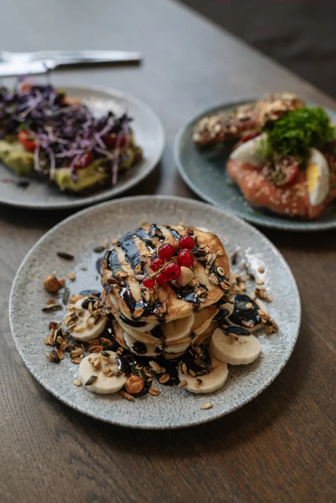 Ein Teller mit Pfannkuchen von der Naturbackstube Wiesender, belegt mit Bananenscheiben, roten Beeren, Samen, Nüssen und Schokoladensoße, steht auf einem Tisch, während im Hintergrund zwei Teller mit herzhaften Speisen verschwimmen.