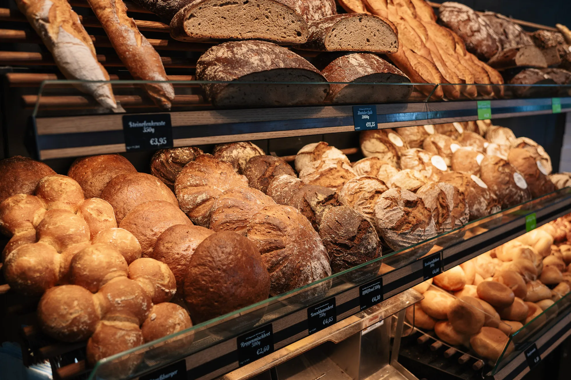 In der Bäckerei Wiesender in Pfaffenhofen werden verschiedene Brotsorten wie Baguettes, runde Brote, knusprige Semmeln und geschnittenes Brot aus der Naturbackstube in Holzregalen ausgestellt.