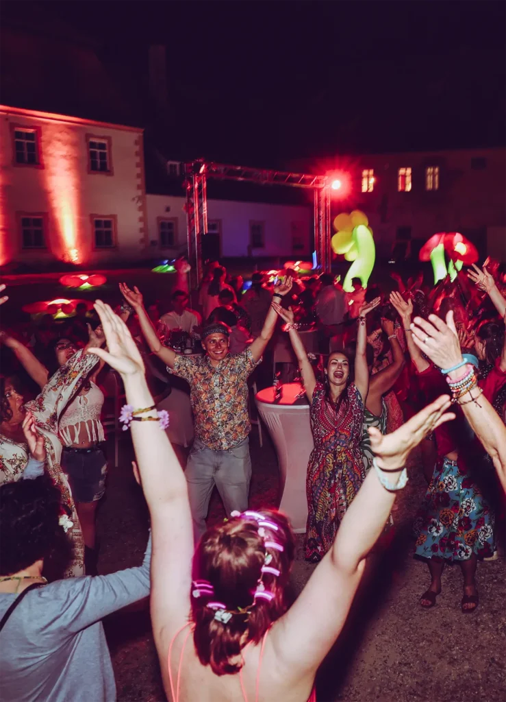 Eine Gruppe von Menschen tanzt nachts unter freiem Himmel in Pfaffenhofen, die Arme erhoben unter buntem Bühnenlicht. Fröhlich und in festlicher, böhmischer Kleidung feiern sie ein ausgelassenes Sommerfest der Naturbackstube Wiesender.