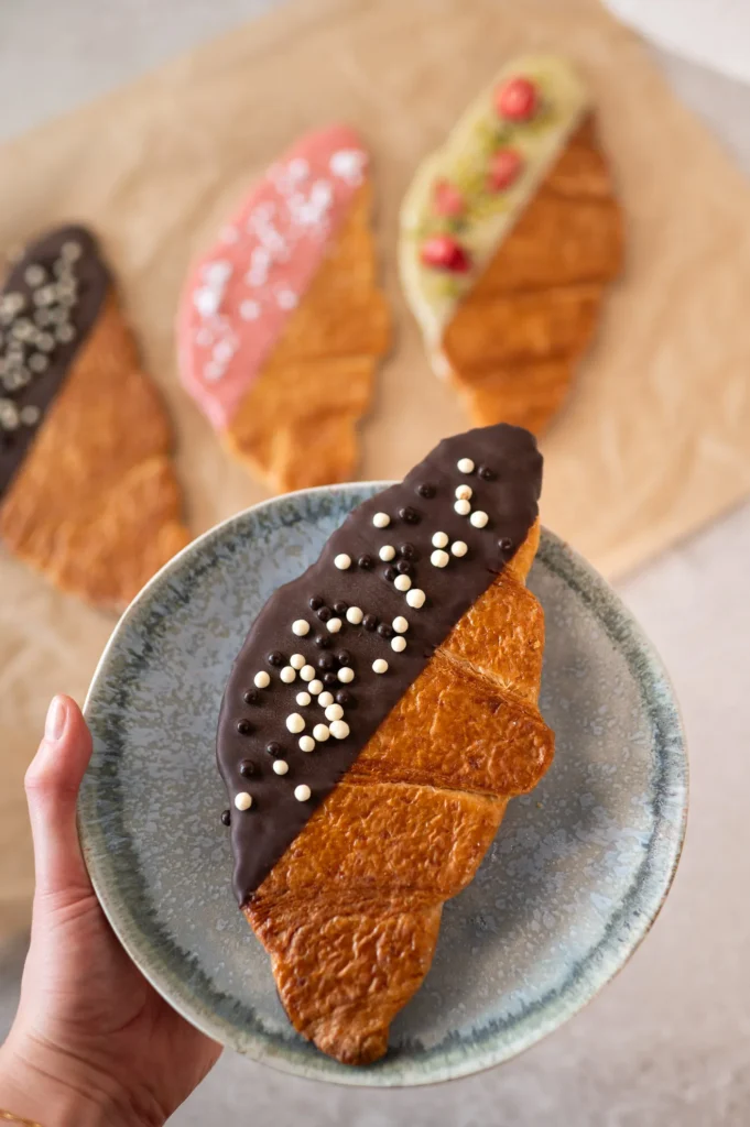 Eine Hand hält einen Teller mit einem Croissant, das halb in Schokolade getaucht und mit weißen und dunklen Schokoladenperlen bestückt ist; im Hintergrund sind drei verzierte Croissants auf Pergamentpapier zu erkennen.
